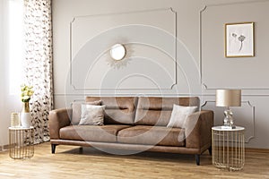 Gold lamp on table next to leather settee in grey living room interior with poster and mirror