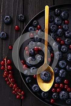 The gold metal spoon, black wood desk surface and fresh natural berries: blueberries and red currant on black dish on a table. top