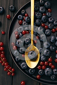 The gold metal spoon, black wood desk surface and fresh natural berries: blueberries and red currant on dish on a table