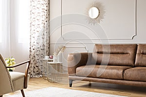 Gold mirror on grey wall with molding above leather settee in flat interior with tables and armchair