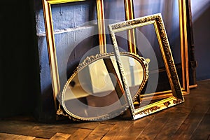 Gold picture frames and a retro oval mirror are arranged in a heap against a backdrop of wood floor and dirty wall. Close-up