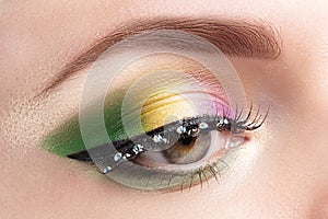 Gold, purple and green Mardi gras makeup. Woman closed eye with bright eyeshadows and rhinestones on false eyelashes closeup