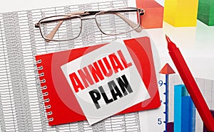 Gold-rimmed glasses, a red pen, color tables and a red notebook with a white card with the text ANNUAL PLAN on the desktop.