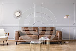 Gold tables in front of leather sofa in grey elegant living room interior with mirror and lamp