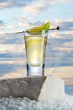 Gold tequila in a glass with slice of lime on a skewer stands on a stone against a beautiful sunset in the desert