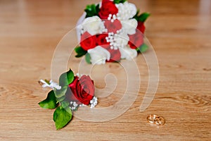 Gold wedding rings, groom`s boutonniere and bride`s bouquet of red and white roses on a wooden background. wedding