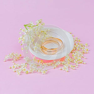 Gold wedding rings in a white seashell with beautiful white elderflower flowers on a pink pastel background.