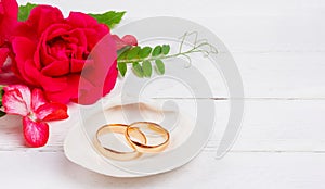 Gold wedding rings in white seashell and red rose flowers on white wooden background