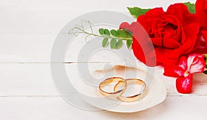 Gold wedding rings in a white seashell and red rose flowers on a white wooden background with copy space.