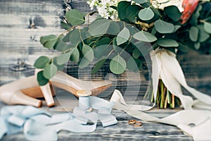 Gold wedding rings on a wooden base with beige bride`s shoes, a spool of ribbon and a bouquet of flowers.