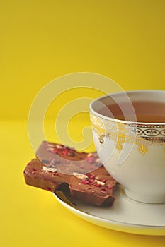 Gold and white porcelain tea cup and saucer with artisan chocolate on bright yellow background with copy space