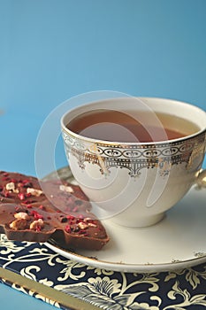 Gold and white porcelain tea cup and saucer with artisan chocolate and open book on blue background with copy space
