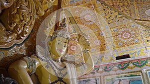 Golden Buddha statue stands in ornate temple. Traditional art adorns walls, ceiling with vibrant colors, patterns