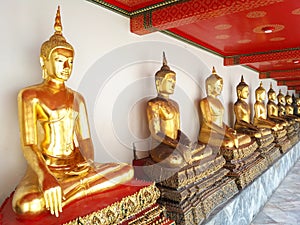 Golden Buddha statues sitting in a meditative pose, in a temple complex with marble floors and a red traditional ceiling in