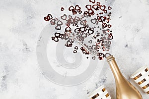 Golden champagne party bottle with confetti in the form of heart and paper cups on bright background