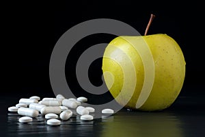 Golden delicious apple and different tablets and capsules on a black background with reflections. Health care, healthy eating and