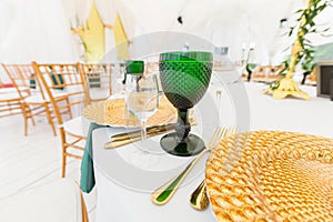 Golden dishes, green wine glasses and napkins. Catering concept. Interior of tent for wedding dinner, ready for guests