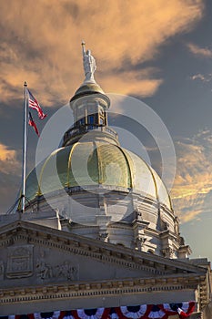 The golden dome of the Georgia Capitol Museum with an American flag and a Georgia State flag with powerful clouds at sunset