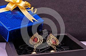 Golden earring studed with red stone in brown box on black background with a blue giftbox. Earing gift concept