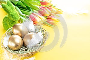 Golden eggs in basket with spring tulips, white feathers on pastel yellow background in Happy Easter decoration. Foil minimalist