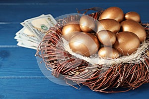 Golden eggs in nest and dollar banknotes on blue wooden table, closeup. Pension concept