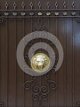 A golden embossed lion's head surrounded by a wrought iron pattern is located on a brown iron fence. Full screen photo
