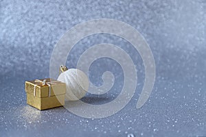 Golden gift box and white Christmas ball ornament on silver sparkling glittery background. Photo with selective focus