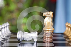 A golden horse chess piece standing on a stack of dollar coins and a silver horse chess piece failing to beg for mercy on a chess