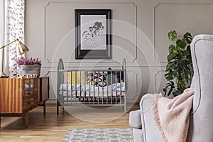 Golden lamp and heather in patterned pot on the vintage cabinet in grey baby room interior with crib and armchair