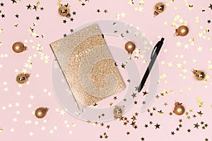 Golden notebook with shiny glitter cover, gold Christmas balls, baubles and confetti stars on pink background