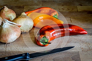 Golden onions and sweet organic pointed peppers with a kitchen knife on a wooden cutting board