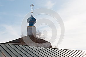 A golden orthodox cross on top of a blue dome on top of the stone white church with a green roof against a blue clear sky