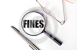 Golden pen and glasses, black magnifying glass with the text FINES on a light background