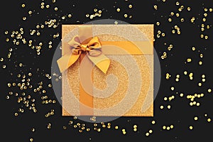 Golden present box with  bow on black background with sequins. Christmas or birthday gift. Celebration and party concept. Flat lay