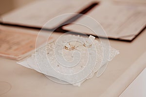 2 golden rings on white wedding pillow. Folder with documents on signing marriage certificate, golden rings closeup. Folder with