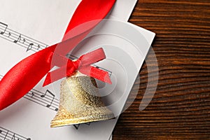Golden shiny bell with red bow and music sheet on wooden table, flat lay. Space for text. Christmas decoration