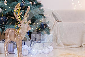 Golden shiny reindeer toy with white baubles, wrapped presents and decorated fir-tree, sofa on blurry background. Winter