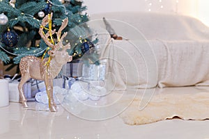 Golden shiny reindeer toy with white baubles, wrapped presents and decorated fir-tree, sofa on blurry background. Winter holidays