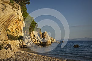 Golden sunlight bathes the rocky cliffs, pine trees on the pebbled shores of the Adriatic Sea in Makarska, Croatia