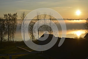 Golden sunrise over the lake in the fog. Smoke over water and silhouettes of small houses and trees