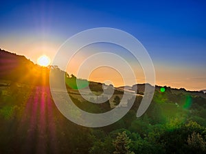 Golden sunrise over trees on the horizon of the hill with strong colorful flares and blue sky