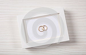 Golden wedding rings on a blank white photo frame, top view, on a white wooden background.