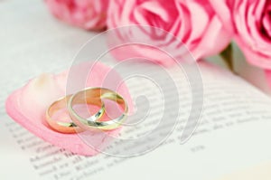 Golden wedding rings on pink rose petal with pink rose flower bouquet on open book with vintage tone