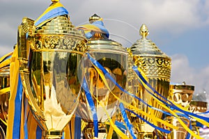 Golden winner's cups with yellow and blue ribbons prepared for awards in a row on the table on sky background