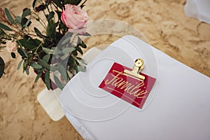 Golden written text familie on red paper with golden clip laying on white cloth. Pink flower and sandy ground in photo
