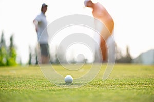 Golf ball approach to the hold on the green. Couple golf player putting golf ball in the background.