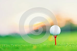 Golf ball on tee at tee off with blur green grass foreground and blur colorful sky with silhouette trees background during sunrise