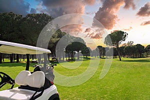 Golf cart in fairway of golf course with green grass field with cloudy sky and trees at sunset