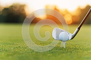 The golf club behind the golf ball on the stand. Against the background of grass and sunset