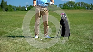 Golf coach holding iron club, preparing to hit ball, sport equipment bag nearby